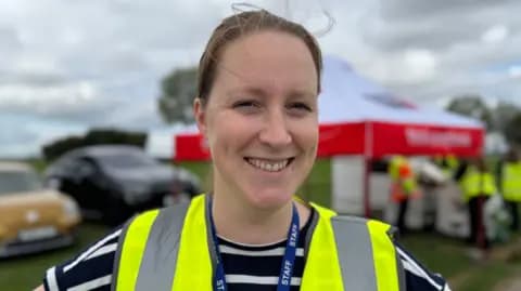 Sharon Edwards/BBC A woman with brown hair, tied back, smiles as she stands in front of a marquee on a showground, with cars parked behind her on a green field. She is wearing a high-vis, yellow vest over a black and white striped top.